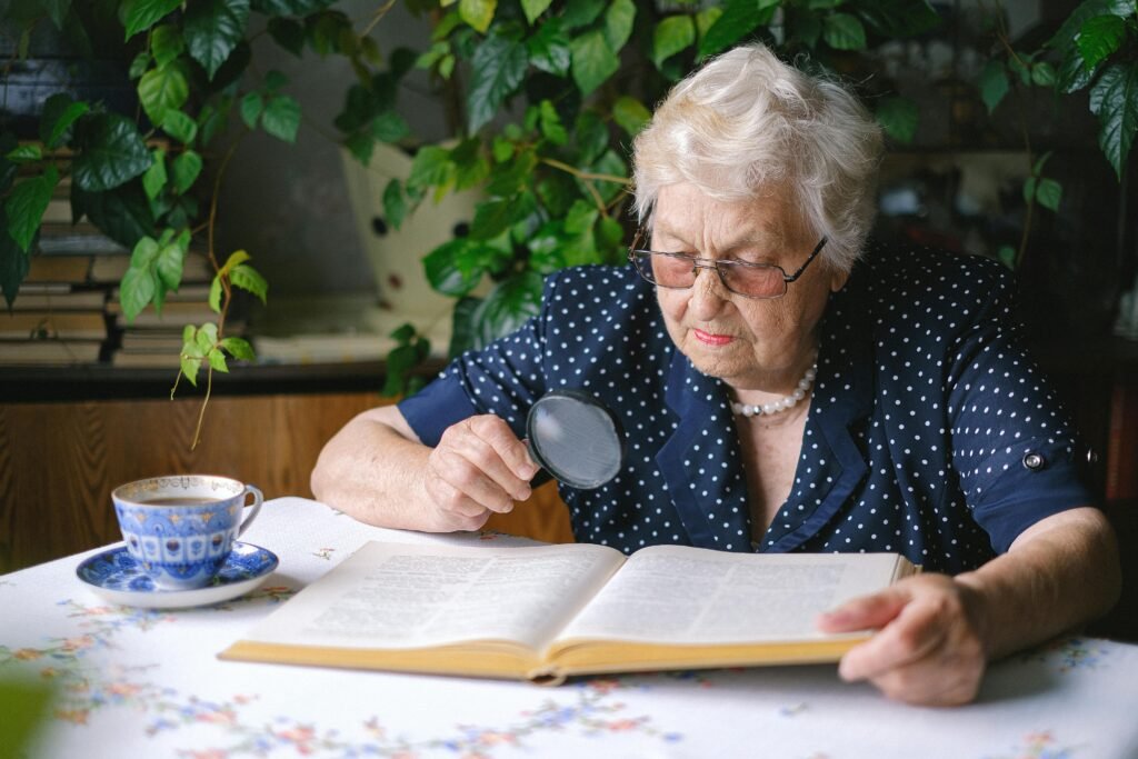 pexels-photo-5231265-5231265 Elderly woman reading a book at home with a magnifying glass, surrounded by plants.